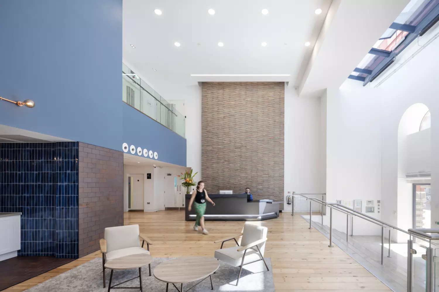 A spacious modern lobby with high ceilings and natural light streaming through large windows. The reception desk sits against a tall brick wall, while the seating area includes two chairs and round tables on a rug. Blue tiled walls and wooden flooring add texture and warmth to the contemporary design.