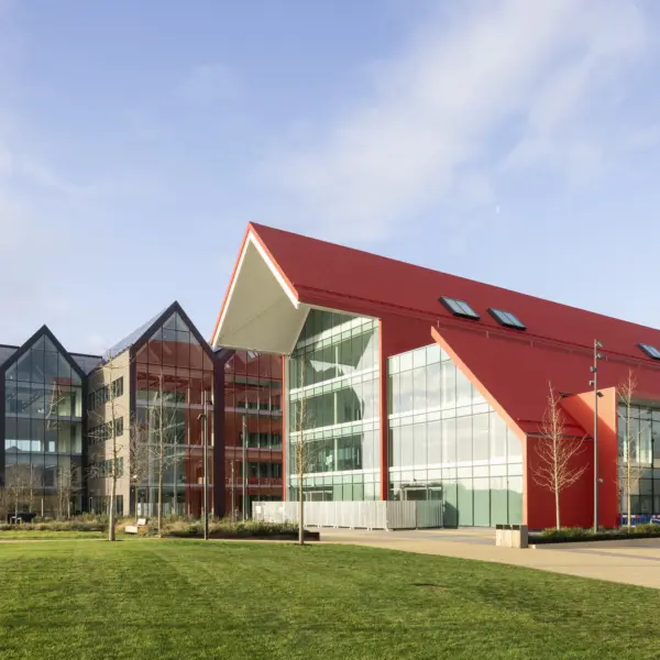 Modern campus-style building with a red pitched roof and large glass façades, connected to a darker grey, multi-gabled glass structure on the left. The buildings sit beside a wide paved walkway and a neatly maintained green lawn in the foreground. Sculptural metal frames holding spherical artworks stand on the grass. The scene is brightly lit by sunlight under a clear blue sky with light clouds, highlighting the clean lines, reflective windows and contemporary architectural design.