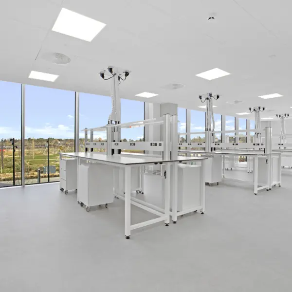Bright, modern laboratory or classroom space with white workbenches arranged in rows on a smooth grey floor. Each bench is fitted with overhead service units suspended from the ceiling, featuring multiple outlets and flexible connections. Large floor-to-ceiling windows line one side of the room, allowing natural light to flood the space and offering views of landscaped grounds outside. The ceiling is fitted with recessed square lighting panels and ventilation fixtures, reinforcing the clean, clinical environment.