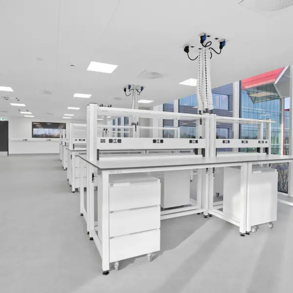 Spacious, modern laboratory with rows of white workbenches fitted with integrated shelving and suspended service units overhead. Each unit features multiple outlets and flexible connections, positioned above the benches for easy access. Large floor-to-ceiling windows run along the right side, filling the room with natural light and revealing views of landscaped grounds and a contemporary red-roofed building outside. The interior has a clean, minimal aesthetic with a grey floor, white ceiling panels with recessed lighting and ventilation fixtures, and additional cabinetry and equipment lining the far wall.