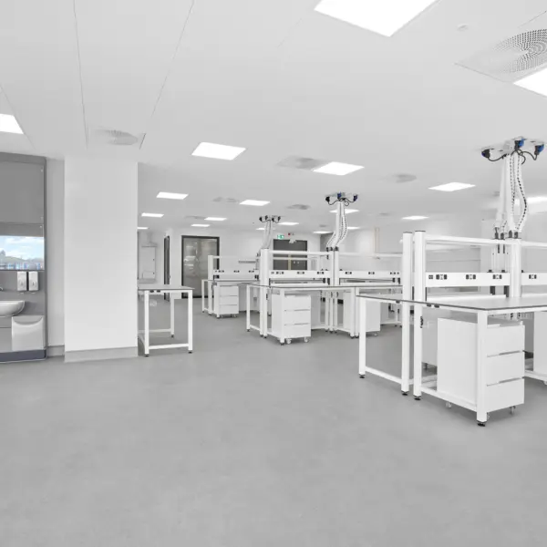 Spacious, modern laboratory fitted with white workbenches arranged across a wide, clean grey floor. Each bench features integrated storage drawers and overhead service units suspended from the ceiling, equipped with flexible hoses and power or data outlets. The ceiling is lined with recessed square light panels and circular ventilation grilles, creating an even, bright illumination throughout the room. On the left, a recessed wash station with a sink, mirror and grey panel detailing sits beside a column, while the rest of the space extends open and uncluttered towards the back, where doors and additional work areas are visible. The overall setting is minimal, clinical and highly organised, with a focus on functionality and clean lines.