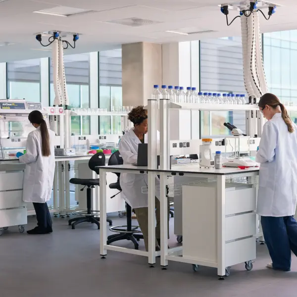 Bright, modern laboratory with large floor-to-ceiling windows, white workstations and scientific equipment. Three researchers in white lab coats work at separate benches, using laboratory instruments and computers. Shelving holds rows of sample bottles and glassware, while ceiling-mounted extraction arms hang above the work areas. The space is clean, open and filled with natural daylight.