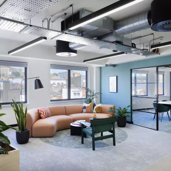 A modern office space featuring a seating area and a meeting room. The seating area includes a semi-circular orange sofa with colourful cushions, a green chair and a small round table. Large windows provide natural light and views of buildings outside. The ceiling has exposed ductwork and modern lighting fixtures. Several potted plants add greenery to the space. The meeting room, visible through glass walls, contains a table with chairs around it.