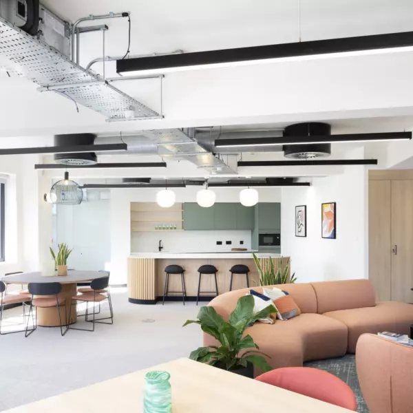 A modern open-plan office featuring industrial-style exposed ceiling ducts and sleek black linear light fixtures. The space includes a kitchen area with light wood cabinetry, white countertops and black bar stools. A round dining table with four chairs sits near a large window that floods the room with natural light. The foreground showcases a peach-coloured L-shaped sofa surrounded by green plants and small tables, creating a relaxed lounge area. Framed artwork adds visual interest to the neutral-toned walls.