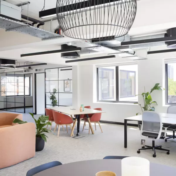 A modern office space featuring a large round light fixture hanging from a ceiling with exposed ducts and electrical conduits. Multiple windows allow natural light to enter. The room includes a round table with candles, an orange couch, a rectangular table with red chairs around it and two desks with office chairs. Several potted plants are placed throughout the space. The overall design is open and airy with a mix of functional and decorative elements.