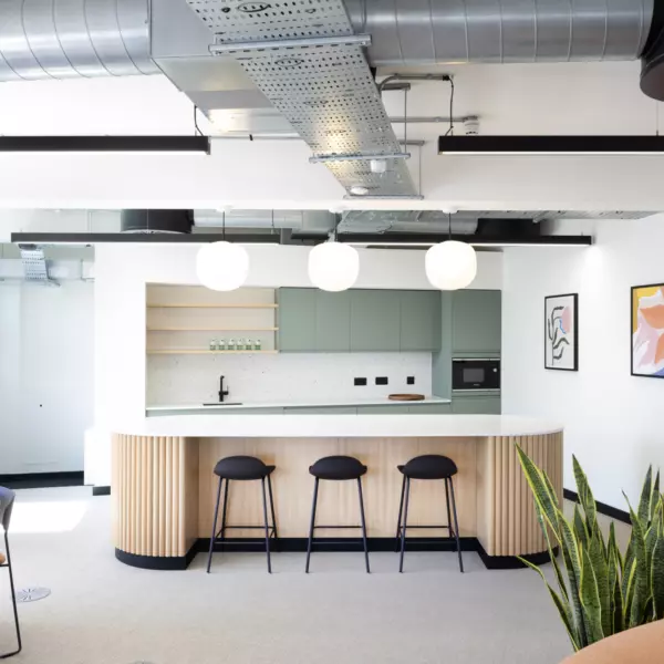 A modern kitchen area with a large island featuring a white countertop and wooden panelled sides, accompanied by three black bar stools. Above the island hang three round pendant lights. The background includes light green cabinets, open shelves with various items, a built-in oven and a sink. The ceiling showcases exposed ductwork and industrial-style lighting fixtures. On the right wall are two framed abstract art pieces. Several plants are placed around the room, including one near a round dining table on the left and another beside a brown sofa on the right.