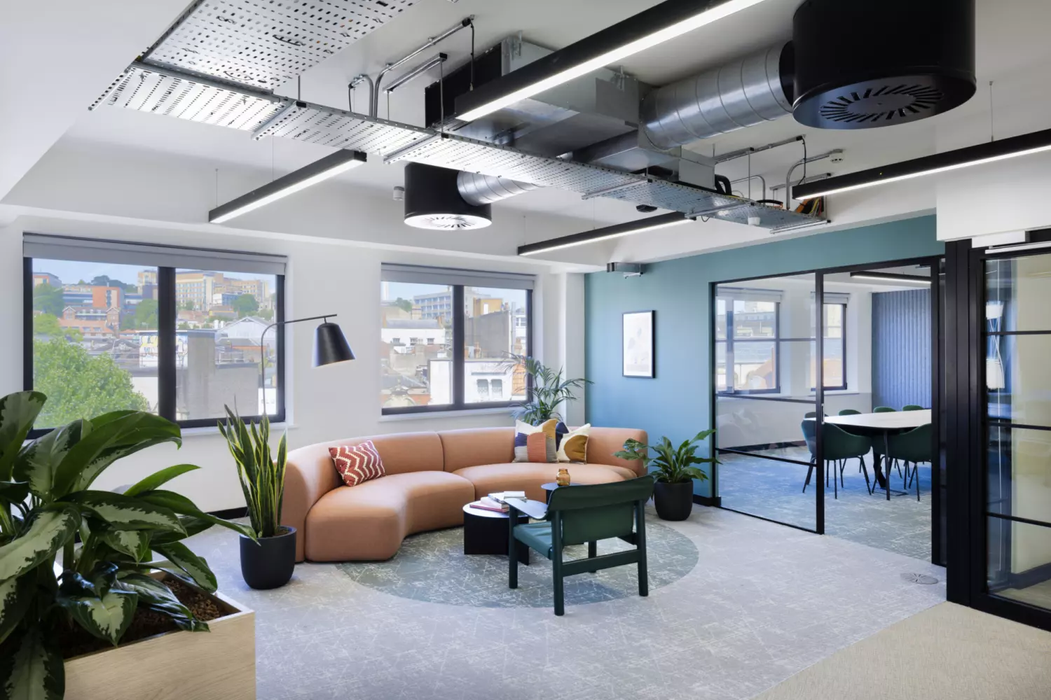 A modern office space featuring a seating area and a meeting room. The seating area includes a semi-circular orange sofa with colourful cushions, a green chair and a small round table. Large windows provide natural light and views of buildings outside. The ceiling has exposed ductwork and modern lighting fixtures. Several potted plants add greenery to the space. The meeting room, visible through glass walls, contains a table with chairs around it.