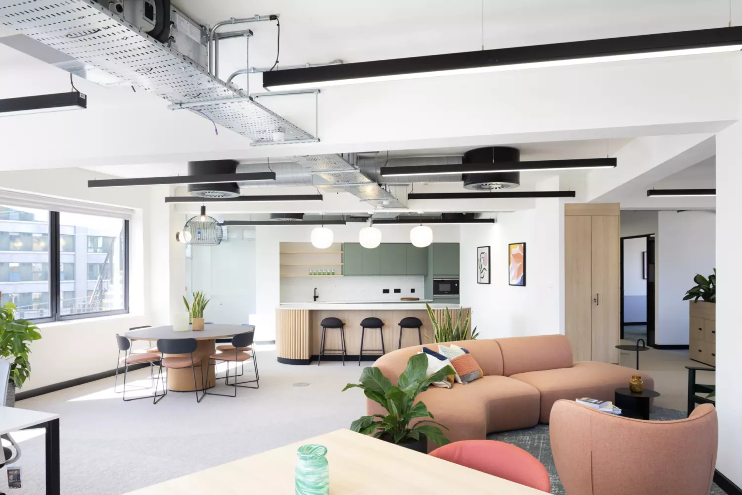 A modern open-plan office featuring industrial-style exposed ceiling ducts and sleek black linear light fixtures. The space includes a kitchen area with light wood cabinetry, white countertops and black bar stools. A round dining table with four chairs sits near a large window that floods the room with natural light. The foreground showcases a peach-coloured L-shaped sofa surrounded by green plants and small tables, creating a relaxed lounge area. Framed artwork adds visual interest to the neutral-toned walls.