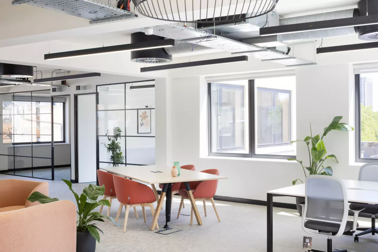 A modern office space featuring a large round light fixture hanging from a ceiling with exposed ducts and electrical conduits. Multiple windows allow natural light to enter. The room includes a round table with candles, an orange couch, a rectangular table with red chairs around it and two desks with office chairs. Several potted plants are placed throughout the space. The overall design is open and airy with a mix of functional and decorative elements.