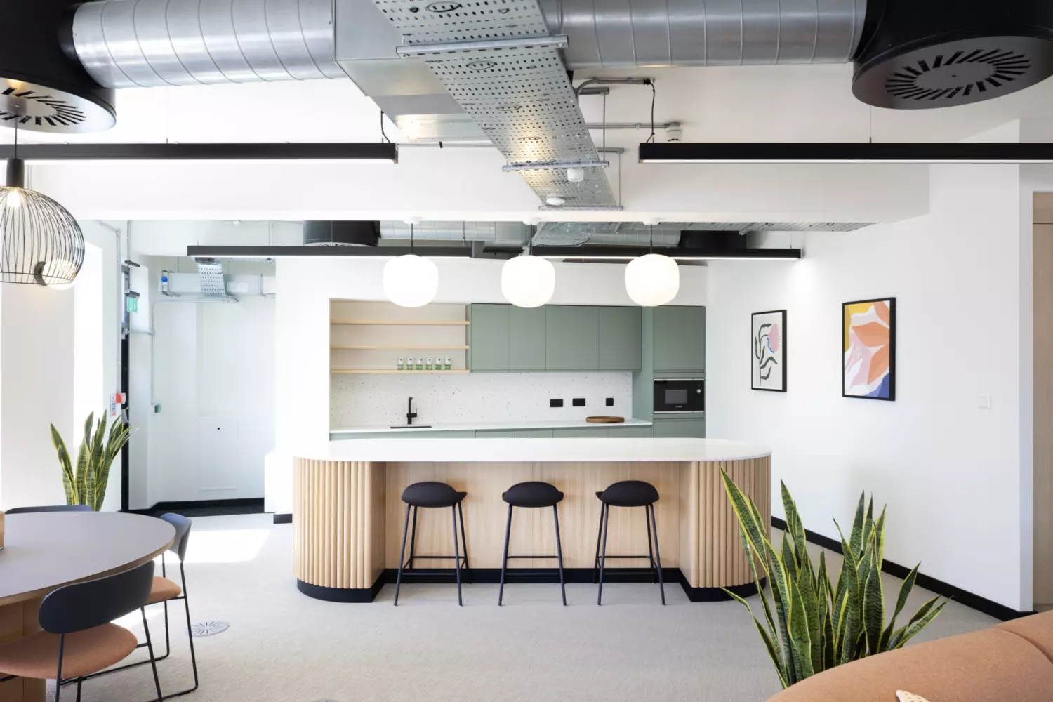 A modern kitchen area with a large island featuring a white countertop and wooden panelled sides, accompanied by three black bar stools. Above the island hang three round pendant lights. The background includes light green cabinets, open shelves with various items, a built-in oven and a sink. The ceiling showcases exposed ductwork and industrial-style lighting fixtures. On the right wall are two framed abstract art pieces. Several plants are placed around the room, including one near a round dining table on the left and another beside a brown sofa on the right.