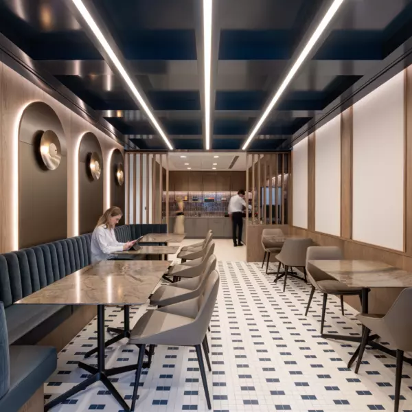 A modern, well-lit office cafe area featuring a long row of tables and chairs. The design includes wooden finishes, dark blue ceiling panels with integrated lighting and tiled flooring. Decorative arches with circular light fixtures line the walls, adding visual interest. At the far end, a counter area is visible, contributing to the space’s functional layout.