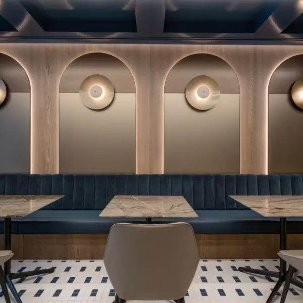 A modern, stylish office cafe style area featuring a wall with three arched niches, each housing a circular light fixture. A long upholstered bench with blue cushioning runs along the wall. In front of the bench are two marble-topped tables, each surrounded by four beige chairs. The floor is finished with patterned tiles in white and blue, adding a decorative touch to the space.