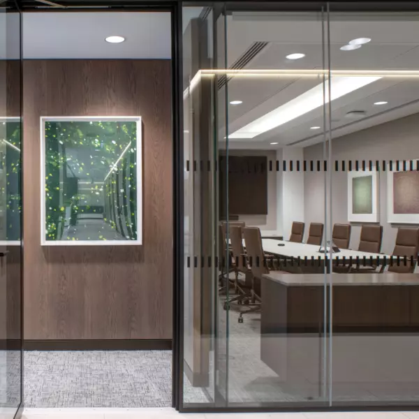 A modern office space featuring a glass-walled conference room with a long table and multiple chairs. The room is decorated with framed artwork on the walls, adding a refined touch. Through the glass, a hallway or entrance area is visible, showcasing another framed piece on a wooden wall, contributing to the cohesive and contemporary design.
