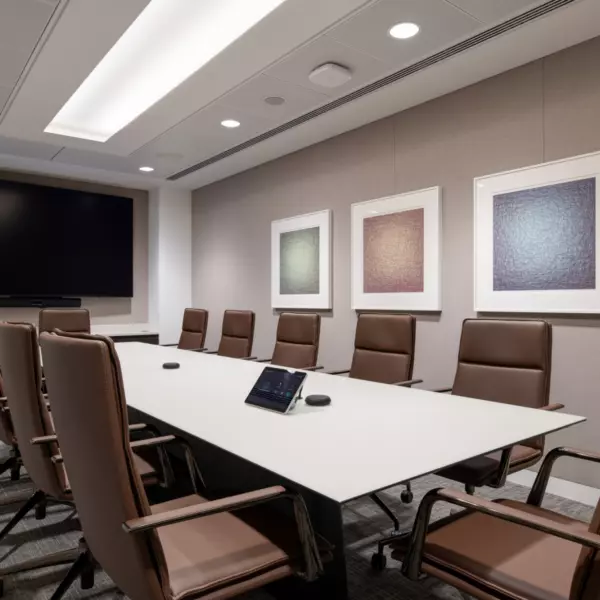 A modern conference room with a long white table surrounded by ten brown leather chairs on wheels. The table holds two small black devices, likely for conferencing or room controls. The back wall is decorated with three framed abstract artworks in muted tones of green, beige and blue. A large flat-screen television is mounted on the far wall. The ceiling features recessed lighting and an illuminated central panel, contributing to the room’s clean and professional aesthetic.