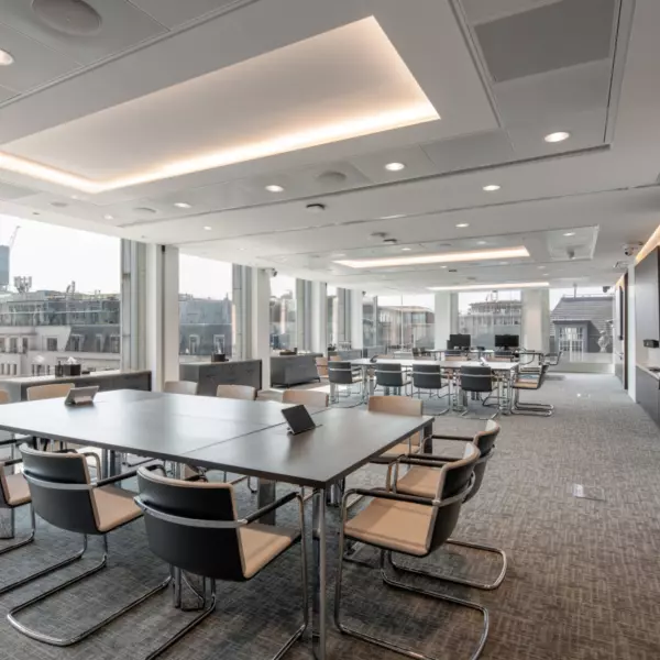 A modern conference room with large windows that allow natural light to fill the space. The room contains two large rectangular tables surrounded by chairs, with additional seating visible in the background. The ceiling features recessed lighting and a flat-screen television is mounted on the right-hand wall. The design is sleek and professional, ideal for meetings or presentations.