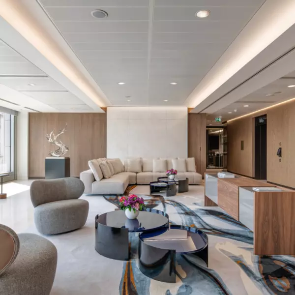 A modern office lobby with a large light-coloured sectional sofa set against a wooden accent wall featuring a central marble panel. The seating area includes round grey upholstered chairs and black and blue circular coffee tables topped with floral arrangements. The floor is covered with an abstract-patterned rug in shades of blue, grey and beige. Large windows on the left allow natural light to fill the space, while the right side has two lifts framed by wooden panelling. The ceiling features recessed lighting and sleek linear fixtures that enhance the contemporary design.