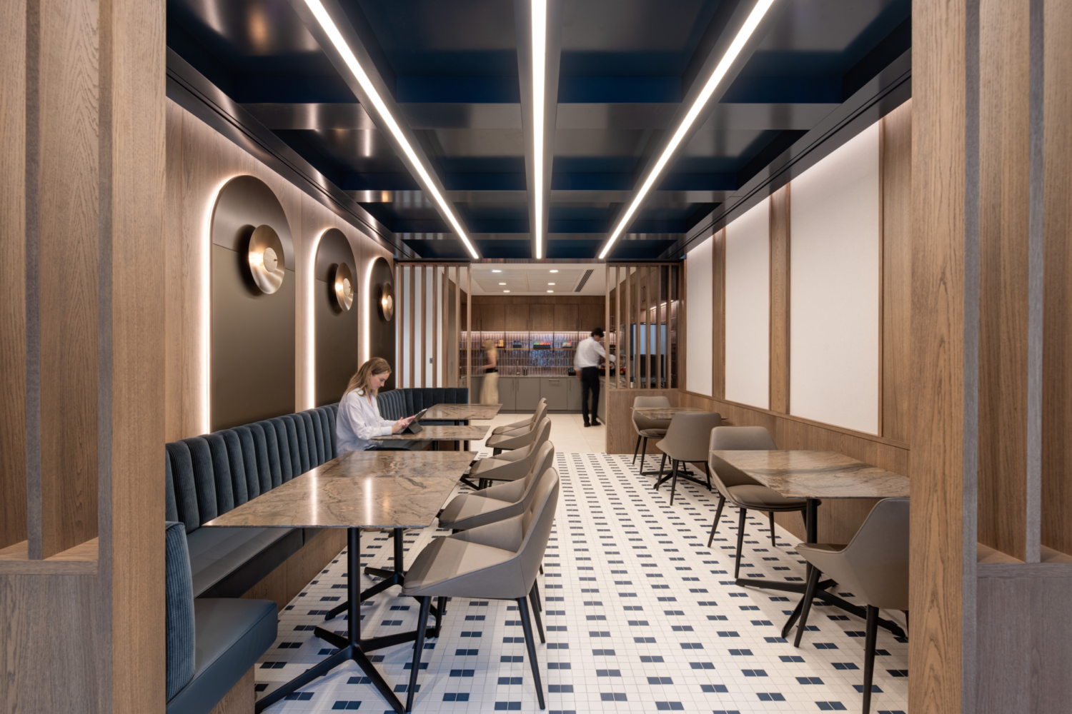 A modern, well-lit office cafe area featuring a long row of tables and chairs. The design includes wooden finishes, dark blue ceiling panels with integrated lighting and tiled flooring. Decorative arches with circular light fixtures line the walls, adding visual interest. At the far end, a counter area is visible, contributing to the space’s functional layout.
