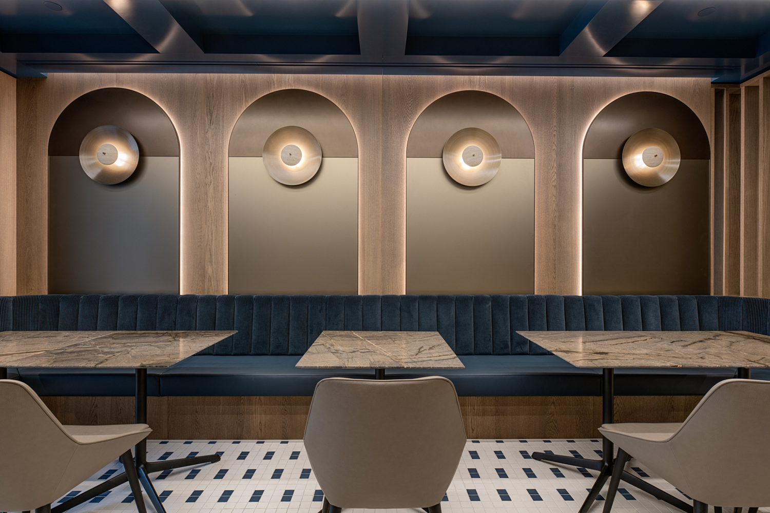 A modern, stylish office cafe style area featuring a wall with three arched niches, each housing a circular light fixture. A long upholstered bench with blue cushioning runs along the wall. In front of the bench are two marble-topped tables, each surrounded by four beige chairs. The floor is finished with patterned tiles in white and blue, adding a decorative touch to the space.