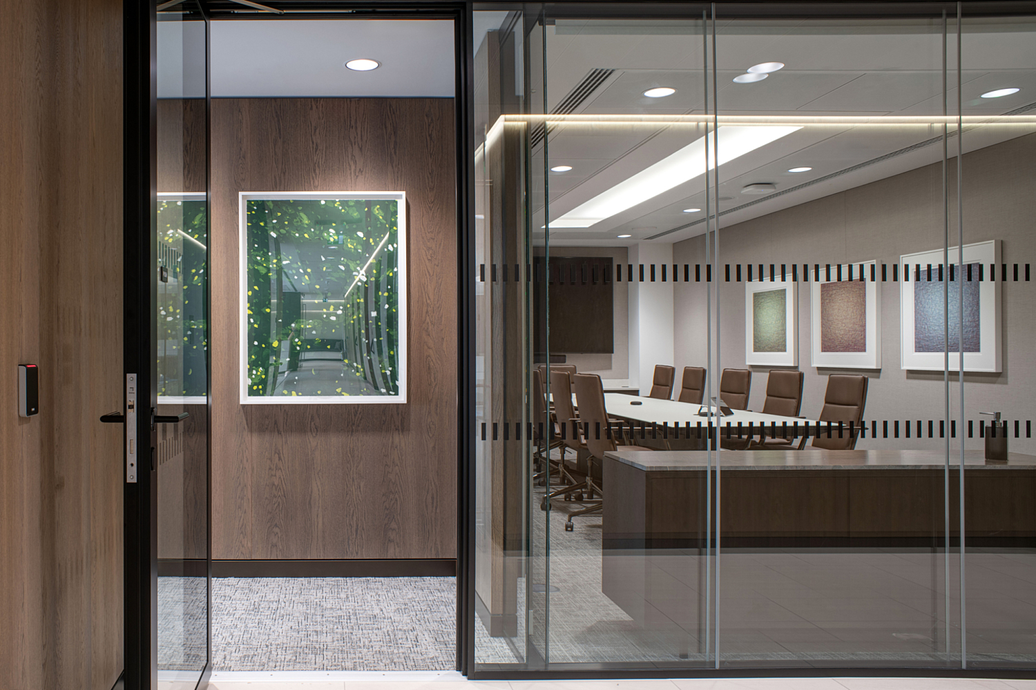 A modern office space featuring a glass-walled conference room with a long table and multiple chairs. The room is decorated with framed artwork on the walls, adding a refined touch. Through the glass, a hallway or entrance area is visible, showcasing another framed piece on a wooden wall, contributing to the cohesive and contemporary design.