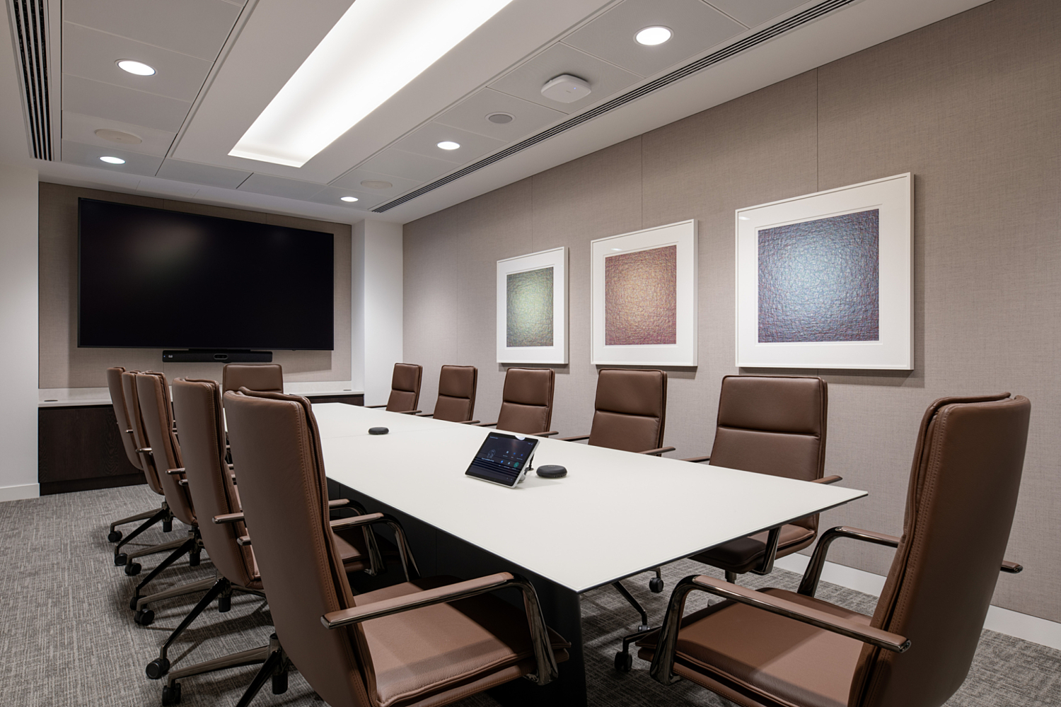 A modern conference room with a long white table surrounded by ten brown leather chairs on wheels. The table holds two small black devices, likely for conferencing or room controls. The back wall is decorated with three framed abstract artworks in muted tones of green, beige and blue. A large flat-screen television is mounted on the far wall. The ceiling features recessed lighting and an illuminated central panel, contributing to the room’s clean and professional aesthetic.