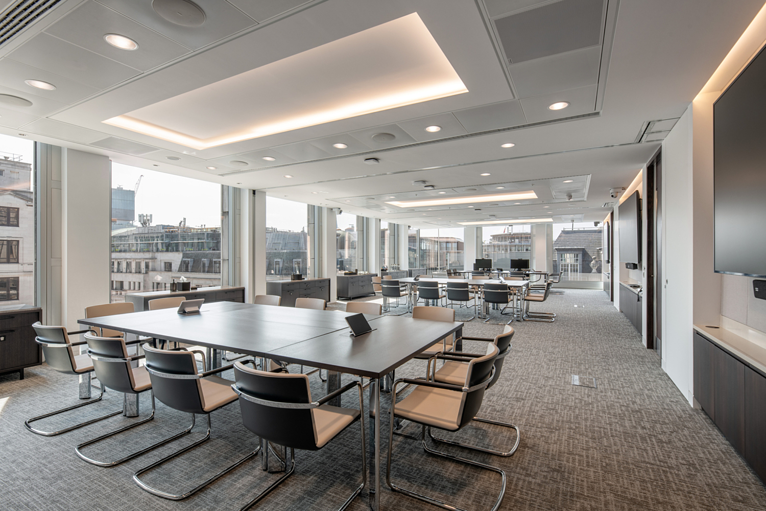 A modern conference room with large windows that allow natural light to fill the space. The room contains two large rectangular tables surrounded by chairs, with additional seating visible in the background. The ceiling features recessed lighting and a flat-screen television is mounted on the right-hand wall. The design is sleek and professional, ideal for meetings or presentations.