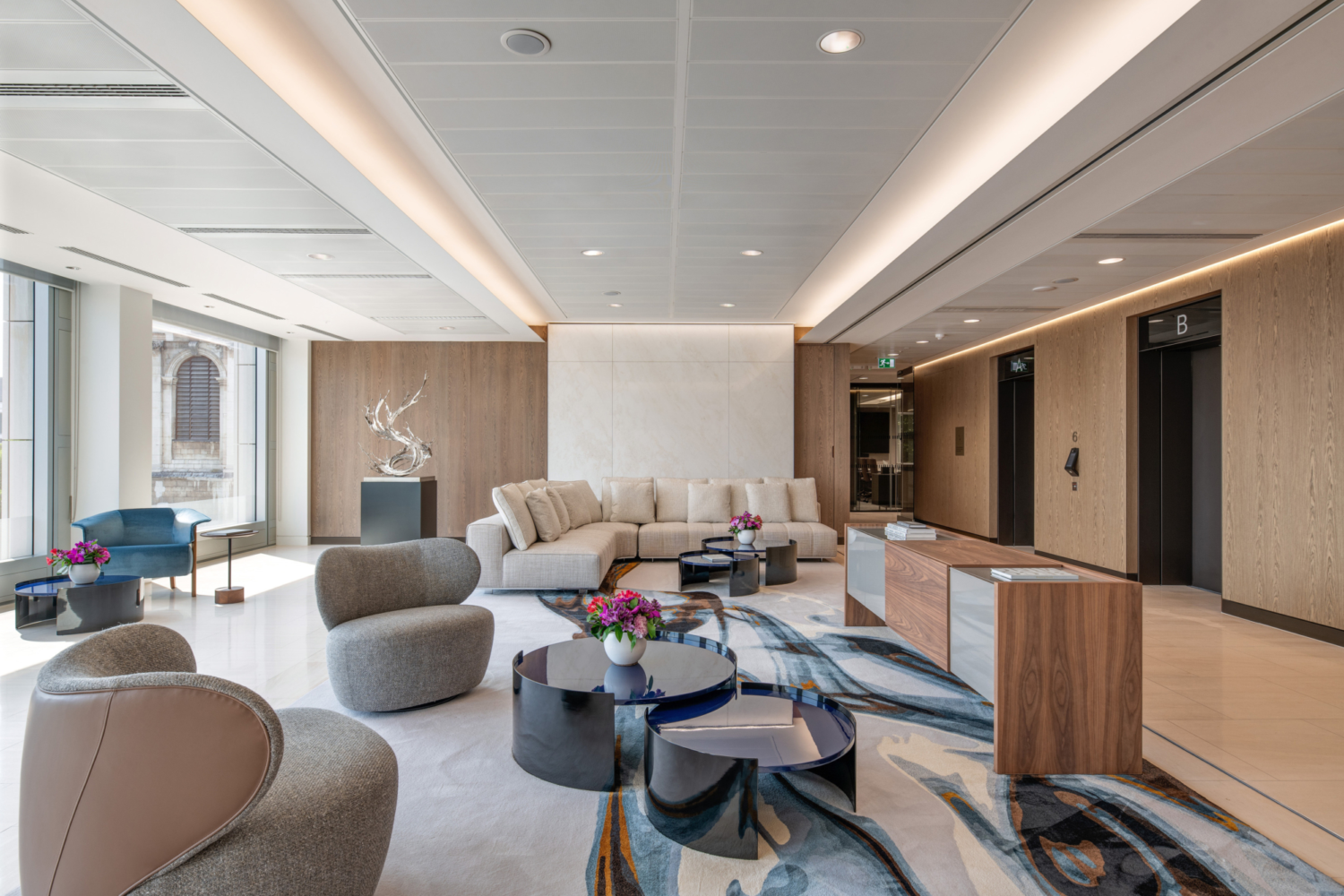 A modern office lobby with a large light-coloured sectional sofa set against a wooden accent wall featuring a central marble panel. The seating area includes round grey upholstered chairs and black and blue circular coffee tables topped with floral arrangements. The floor is covered with an abstract-patterned rug in shades of blue, grey and beige. Large windows on the left allow natural light to fill the space, while the right side has two lifts framed by wooden panelling. The ceiling features recessed lighting and sleek linear fixtures that enhance the contemporary design.