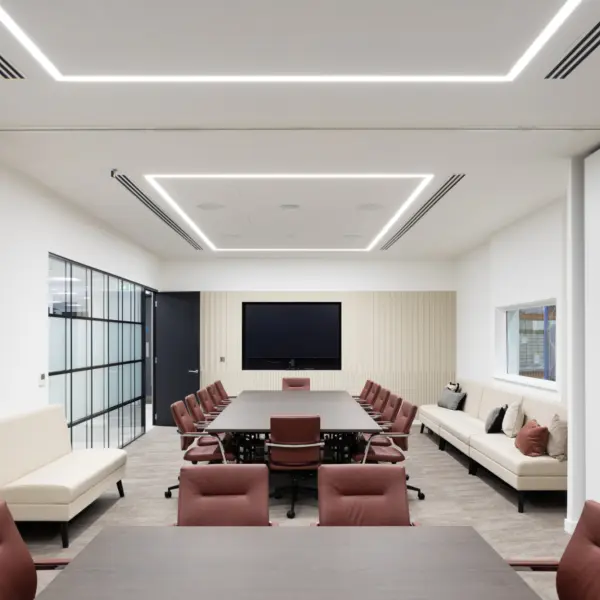 A modern conference room with a large central meeting table surrounded by brown upholstered chairs on castors. At the far end of the room, a wide screen is mounted on a ribbed feature wall. Built‑in sofas with neutral cushions run along both side walls. The room has white walls, a suspended ceiling with integrated rectangular LED lighting, and a glass partition on the left that leads to an adjacent corridor. Sliding partition panels frame the space, the flooring is a mix of carpet and hard surfaces in muted tones.