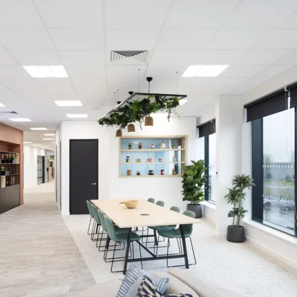 A bright office breakout area with a long light‑wood table and green chairs positioned near large windows with black roller blinds. Above the table hangs a feature light with plants and wicker shades. A shelving unit on the far wall displays assorted jars and containers, with potted plants placed below it. To the left, a corridor leads further into the office and includes a living wall with illuminated signage. The space has white walls, a light carpeted floor and a suspended ceiling with recessed lighting.