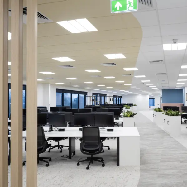 A spacious open‑plan office with rows of white desks and black monitors arranged in clusters. Each desk grouping is paired with black task chairs, and low planters divide sections of the workspace. The ceiling features a mix of white and pale yellow panels with recessed lighting. Large windows line the left side of the room, allowing natural light into the space. In the foreground, vertical wooden slats partially frame the view. The flooring combines carpet and a lighter walkway surface, leading toward additional work areas in the background. A green emergency exit sign is visible on the ceiling.