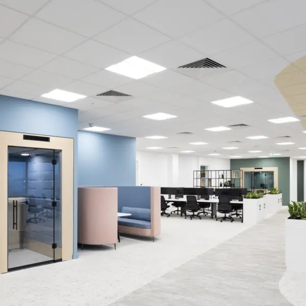 A modern open‑plan office with a light, neutral colour scheme. In the foreground, there is a small enclosed meeting pod with a glass door set within a pale wood frame against a blue wall. Next to it are two high‑backed upholstered booths in soft pastel colours. Beyond this area are clusters of white desks with black office chairs, divided by low planters. The space has a suspended ceiling with recessed lighting, a mix of carpeted and hard flooring and varied wall colours including blue, white and green. The layout is spacious with wide walkways and contemporary finishes throughout.
