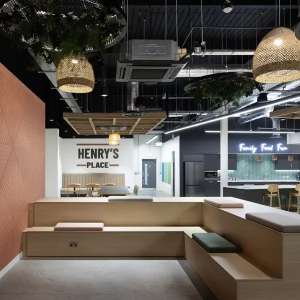 A contemporary breakout space featuring tiered wooden seating with upholstered cushions in muted colours. To the left, a large patterned wall panel frames a mounted screen. The ceiling has exposed ductwork with hanging woven pendant lights and circular planters containing greenery. In the distance, a sign reading “Henry’s Place” is mounted above a small dining area with wooden tables and chairs. Further back, an open kitchen area with a green tiled island and a neon sign reading “Family Food Fun” is visible. The space combines warm timber finishes with industrial ceiling elements and soft seating.