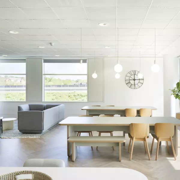 A spacious office lounge area with large windows that allow abundant natural light. The ceiling has a grid layout with recessed lighting fixtures. The flooring is laid in a herringbone pattern, adding visual texture. Furniture includes a long wooden table with matching chairs and benches, as well as a grey sofa set arranged near a wall-mounted television. A minimalist wall clock and a leafy potted plant contribute to the clean, functional aesthetic.