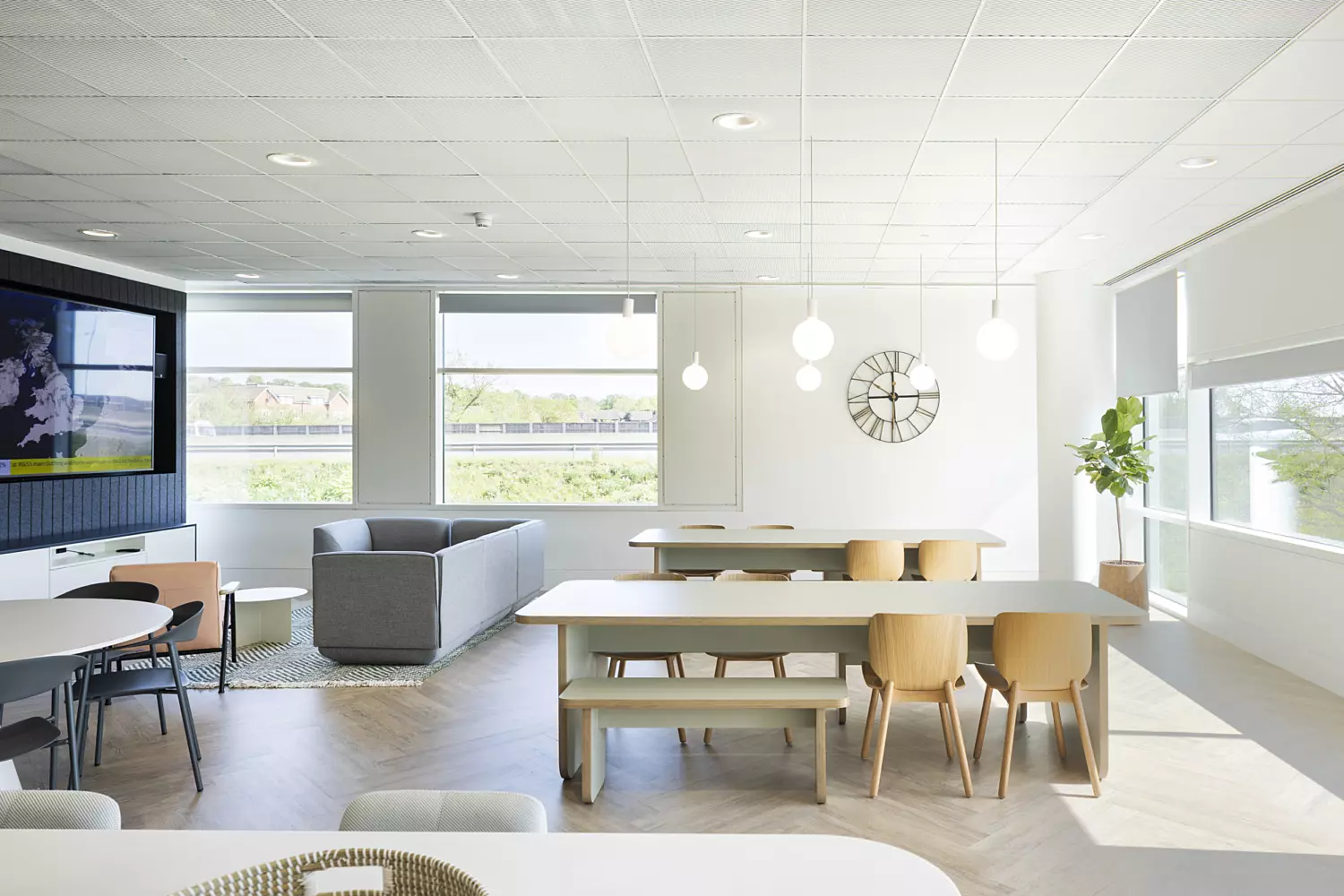 A spacious office lounge area with large windows that allow abundant natural light. The ceiling has a grid layout with recessed lighting fixtures. The flooring is laid in a herringbone pattern, adding visual texture. Furniture includes a long wooden table with matching chairs and benches, as well as a grey sofa set arranged near a wall-mounted television. A minimalist wall clock and a leafy potted plant contribute to the clean, functional aesthetic.
