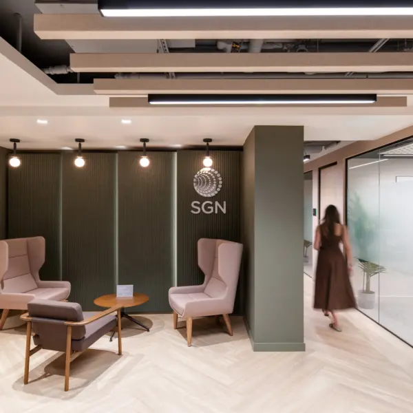 Modern office reception and breakout area featuring lounge seating arranged around a small round table. A textured feature wall displays the SGN logo, while acoustic ceiling panels and pendant lighting create a warm, contemporary atmosphere. Glass-fronted meeting rooms line the corridor, and a person walks through the space towards the offices beyond.