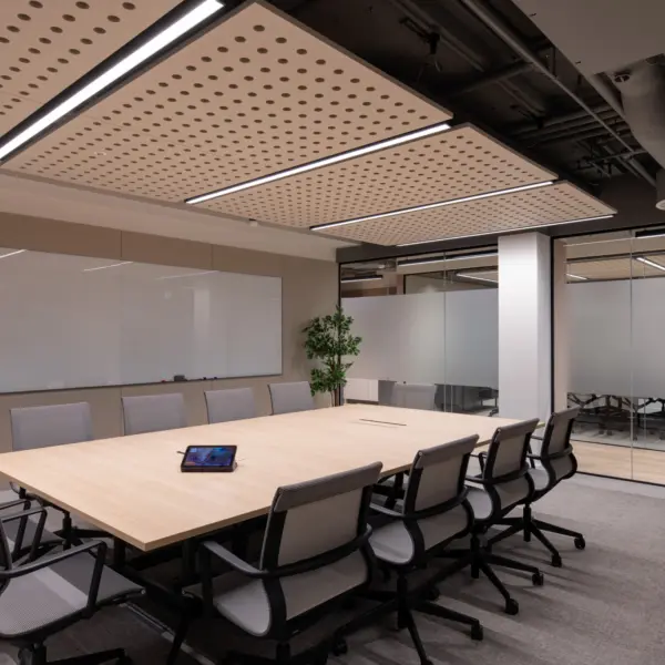 Modern meeting room featuring a large boardroom table surrounded by ergonomic task chairs. A full-width whiteboard spans one wall, while glazed partitions provide views into adjacent meeting spaces. Acoustic ceiling panels and suspended lighting create a comfortable environment for collaboration, with potted plants adding greenery to the contemporary workspace.