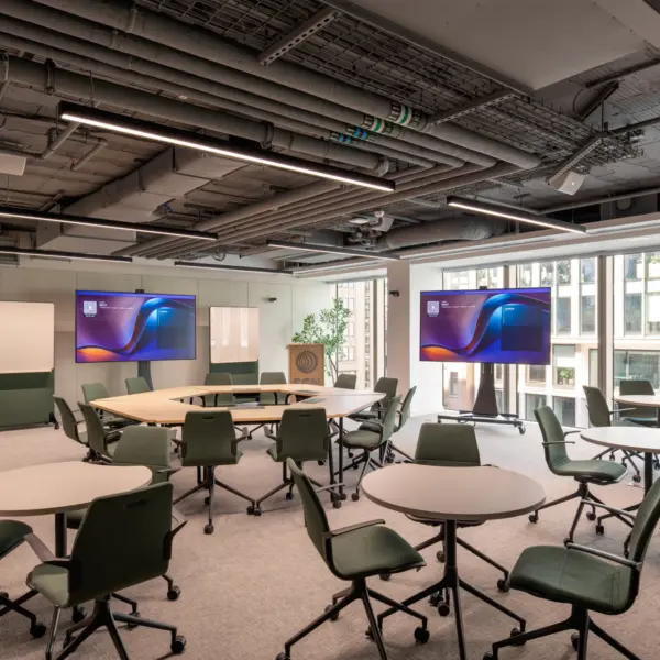Flexible training and collaboration space with movable tables and green task chairs arranged for group work. Two large presentation screens display branded content, while mobile whiteboards provide additional space for workshops and meetings. Floor-to-ceiling windows bring natural light into the room, and exposed ceiling services with contemporary lighting create a modern workplace environment designed for learning and collaboration.