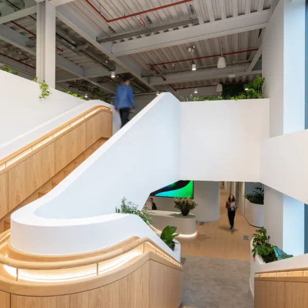 A modern stairwell with white balustrades and timber accents, featuring integrated lighting along the handrails. The staircase splits across multiple levels, with planted areas at the landings. Exposed ceiling services run above, and large windows on the right bring natural light into the space. People are visible moving through the area, giving a sense of scale and activity.