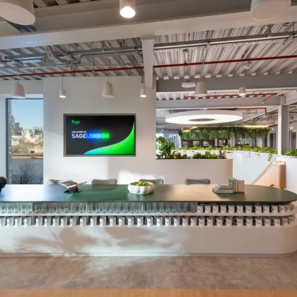 A bright office space with a long green-topped counter in the foreground, backed by neatly arranged rows of cups. A large wall screen is mounted to the left, and daylight enters through a window behind the desk. Beyond the counter, planted partitions frame an open workspace with exposed services, soft lighting and a circular ceiling feature above a greenery-filled area. A staircase with timber treads is visible on the right.