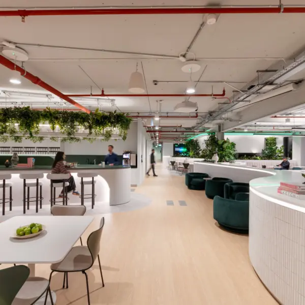 A spacious modern workplace with light wood flooring, white curved counters and abundant greenery. A central bar-style area with stools sits beneath a ceiling feature covered in hanging plants. People are seated at tables and lounge chairs throughout the open space. Overhead pipes and lighting are exposed, and the setting has a bright, contemporary feel.
