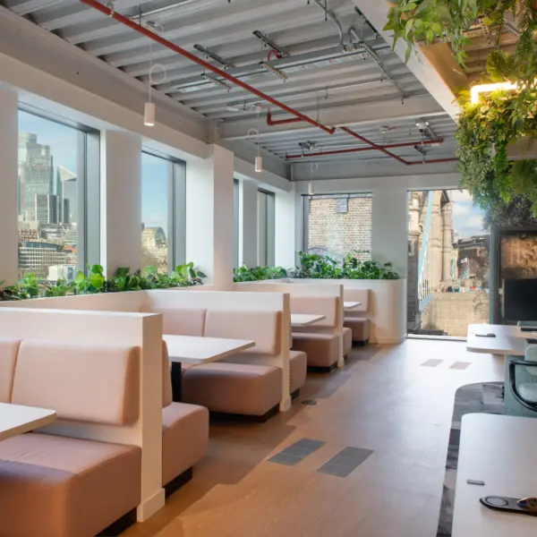 A bright workspace with a row of booth seating arranged beside large windows that offer views of London’s skyline. Light wood flooring runs through the space, with planters separating the booths from the central walkways. The ceiling features exposed services and hanging greenery. Workstations are positioned to the right, creating a mix of focused and informal seating areas.