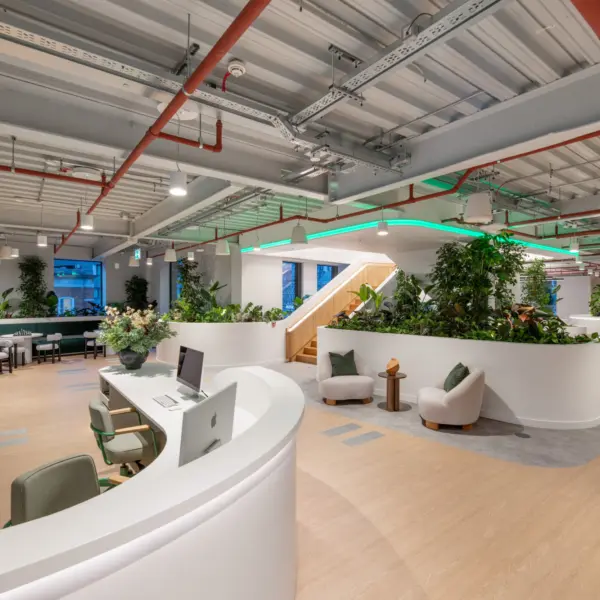 A bright, contemporary workspace with white curved desks, light wood flooring and abundant planting. A large wall screen sits on the left, while a central staircase with timber treads rises behind planted partitions. Lounge chairs, workstations and tables are arranged throughout the open area. Exposed ceiling services and soft integrated lighting complete the modern office setting.