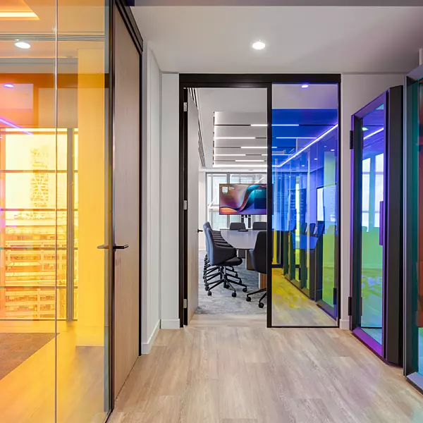 A contemporary office environment featuring glass-walled meeting rooms with tinted panels in a range of colours, including yellow, orange, blue, and green. A hallway with light wooden flooring leads towards a conference room at the far end, which is furnished with multiple chairs arranged around a central table and a large screen mounted on the wall. The ceiling is fitted with recessed lighting and LED strip lights, enhancing the modern and polished aesthetic of the space.