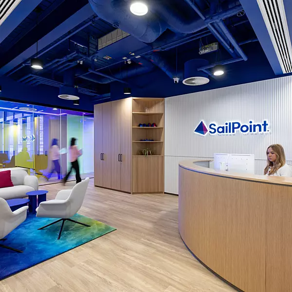 A stylish office reception area featuring a modern receptionist desk positioned on the right, backed by a wall displaying the "SailPoint" logo. The seating area includes contemporary white chairs and a sofa adorned with colourful cushions, complemented by a vibrant, patterned rug that adds visual interest. Glass walls in the background contribute to an open and airy feel, with people visible walking past, enhancing the sense of a dynamic workplace environment.