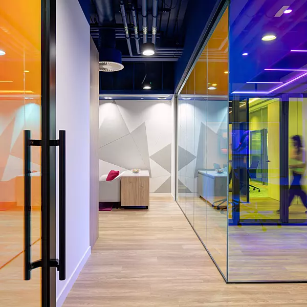 A modern office interior featuring glass walls and doors tinted in bold, vibrant colours including orange, yellow, blue, and purple. The space has a contemporary, open-plan layout with a light wood floor and an exposed ceiling showcasing industrial-style piping and lighting fixtures. In the background, people are walking through the corridor, adding a sense of activity. A small seating area with a sofa and table is visible through one of the coloured glass partitions, contributing to a welcoming and collaborative atmosphere.