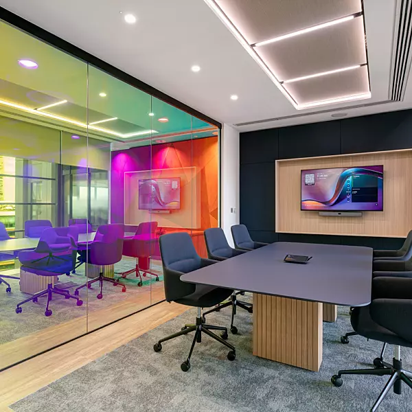 A contemporary office meeting room featuring a large central table surrounded by multiple chairs, designed for collaborative work. A glass wall separates this space from an adjacent meeting area, which is visually enhanced by ambient lighting in vibrant hues of yellow, green, and purple. A large screen is mounted on the wall within the main meeting room, supporting presentations and digital collaboration. The overall design blends functionality with a modern aesthetic.