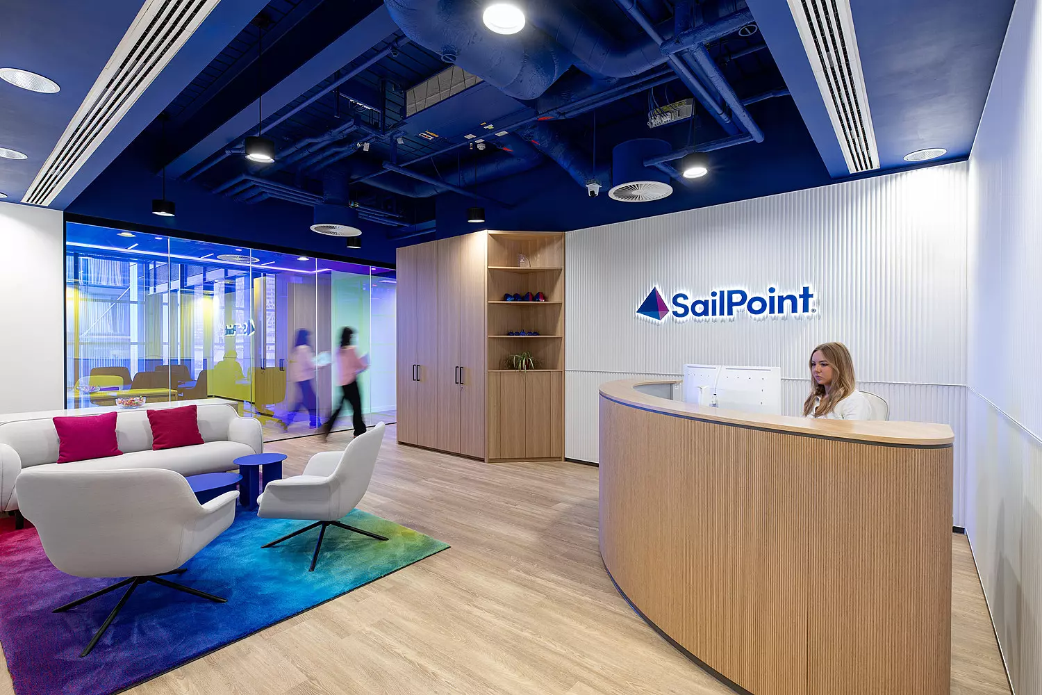 A stylish office reception area featuring a modern receptionist desk positioned on the right, backed by a wall displaying the "SailPoint" logo. The seating area includes contemporary white chairs and a sofa adorned with colourful cushions, complemented by a vibrant, patterned rug that adds visual interest. Glass walls in the background contribute to an open and airy feel, with people visible walking past, enhancing the sense of a dynamic workplace environment.