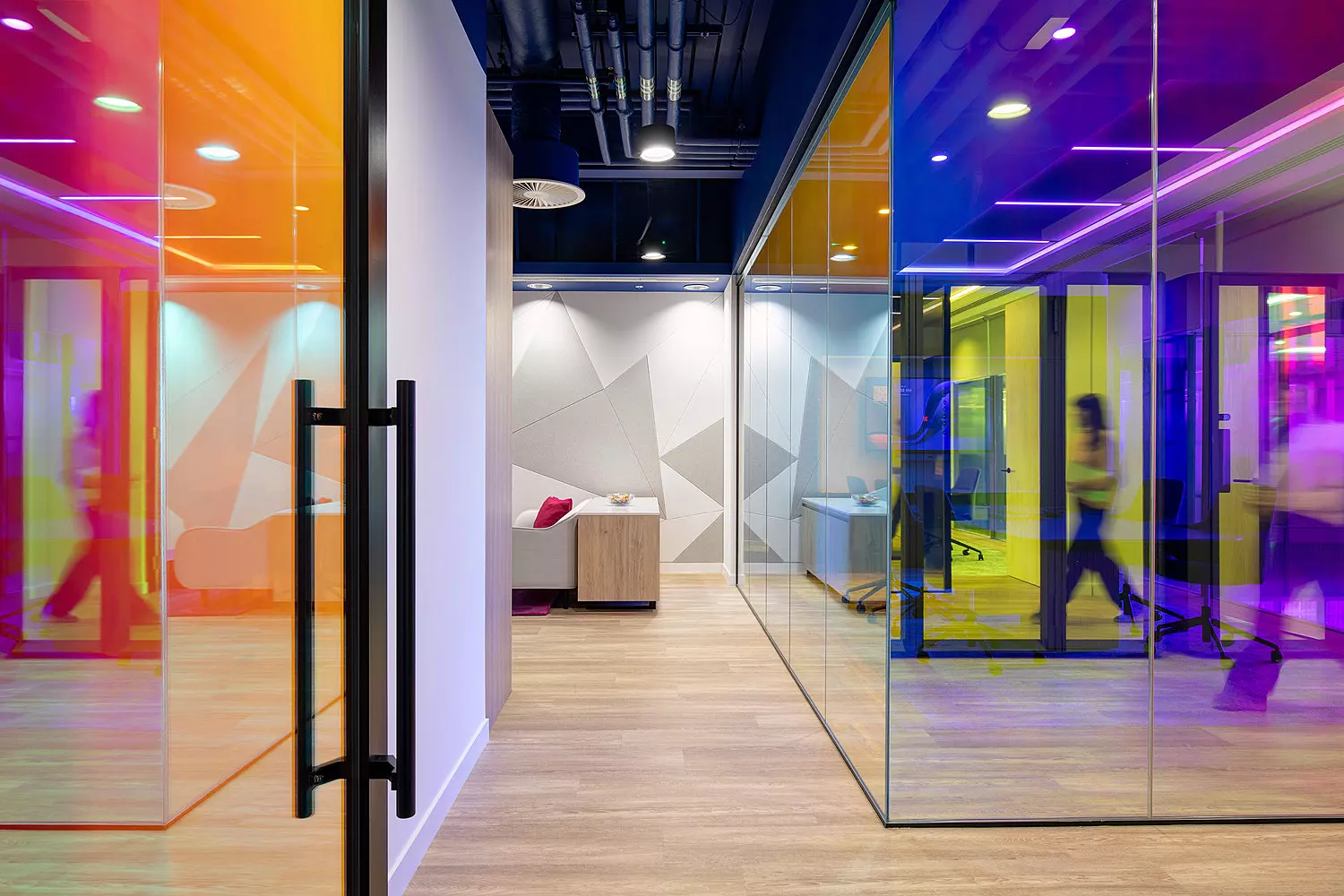 A modern office interior featuring glass walls and doors tinted in bold, vibrant colours including orange, yellow, blue, and purple. The space has a contemporary, open-plan layout with a light wood floor and an exposed ceiling showcasing industrial-style piping and lighting fixtures. In the background, people are walking through the corridor, adding a sense of activity. A small seating area with a sofa and table is visible through one of the coloured glass partitions, contributing to a welcoming and collaborative atmosphere.