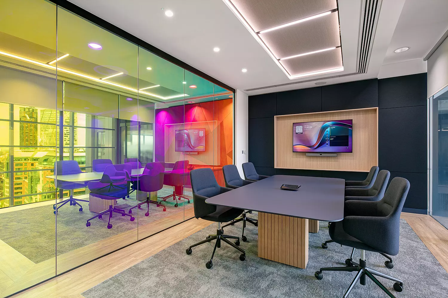 A contemporary office meeting room featuring a large central table surrounded by multiple chairs, designed for collaborative work. A glass wall separates this space from an adjacent meeting area, which is visually enhanced by ambient lighting in vibrant hues of yellow, green, and purple. A large screen is mounted on the wall within the main meeting room, supporting presentations and digital collaboration. The overall design blends functionality with a modern aesthetic.