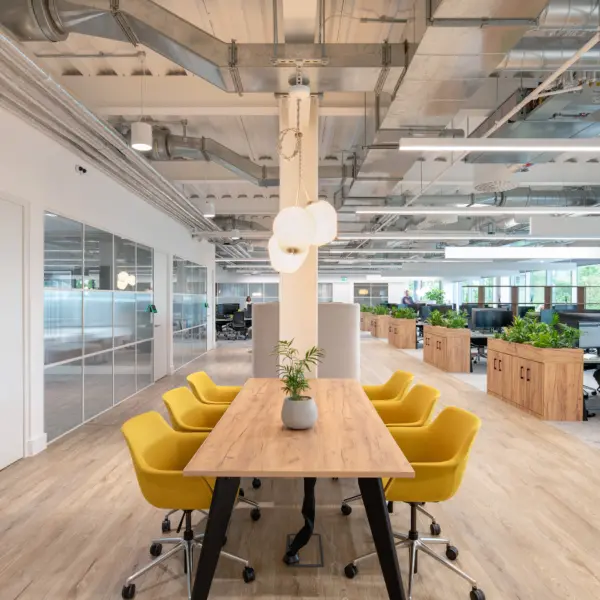 Long, light-filled open-plan office featuring a central wooden meeting table surrounded by six upholstered yellow swivel chairs on wheels. Above the table hang three spherical pendant lights attached to a white structural column. The ceiling is exposed, showing silver ductwork, pipes and suspended linear lighting.  To the left, a row of glass-partitioned meeting rooms with sliding doors runs along the wall, with frosted lower panels and clear upper sections. To the right and extending into the background, rows of desks with computer monitors and office chairs are divided by rectangular wooden planters filled with green plants.  The floor combines light wood in the central walkway with carpeted areas around the workstations. Large windows at the far end allow natural light into the space, and additional potted plants add greenery throughout. The overall setting is modern, clean and airy with a mix of neutral tones and bright accents.