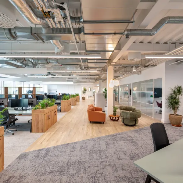Bright, open plan office interior with an exposed ceiling showing silver ductwork and services, white beams and linear lighting. Rows of desks with multiple monitors and task chairs sit to the left, separated by long wooden planters filled with green plants. A central walkway with light wood flooring runs through the space, leading to a soft seating area with upholstered armchairs and a small table. On the right, a glass partition encloses meeting rooms with visible chairs inside, alongside a sideboard, potted plants and wall art. The space combines neutral tones, natural materials and greenery, with carpeted zones around the workstations and a clean, contemporary finish throughout.